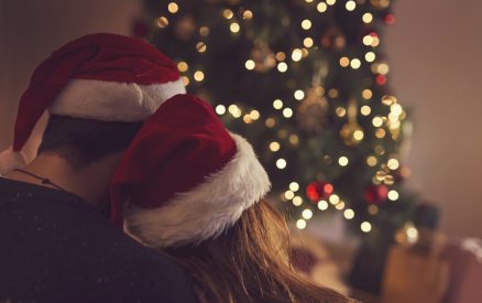 Couple in love sitting next to a Christmas tree, wearing Santa's hats, hugging and looking away from the camera towards the tree. Selective focus on the girl's hat