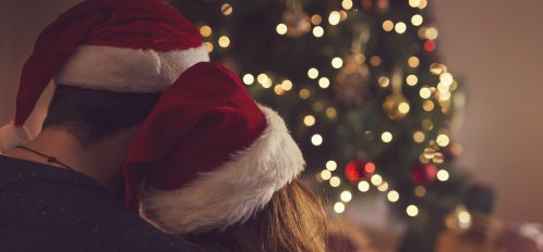 Couple in love sitting next to a Christmas tree, wearing Santa's hats, hugging and looking away from the camera towards the tree. Selective focus on the girl's hat