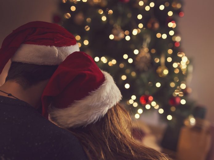 Couple in love sitting next to a Christmas tree, wearing Santa's hats, hugging and looking away from the camera towards the tree. Selective focus on the girl's hat