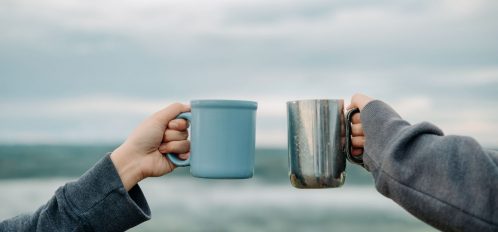 Two people holding coffee cups in front of the ocean.